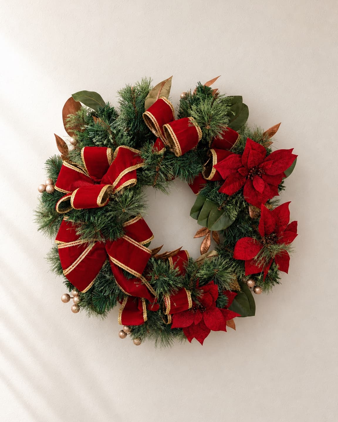 A wreath of fir and magnolia leaves with loops of red velvet ribbon, red poinsettia blooms and gold berries, on a pale plaster wall