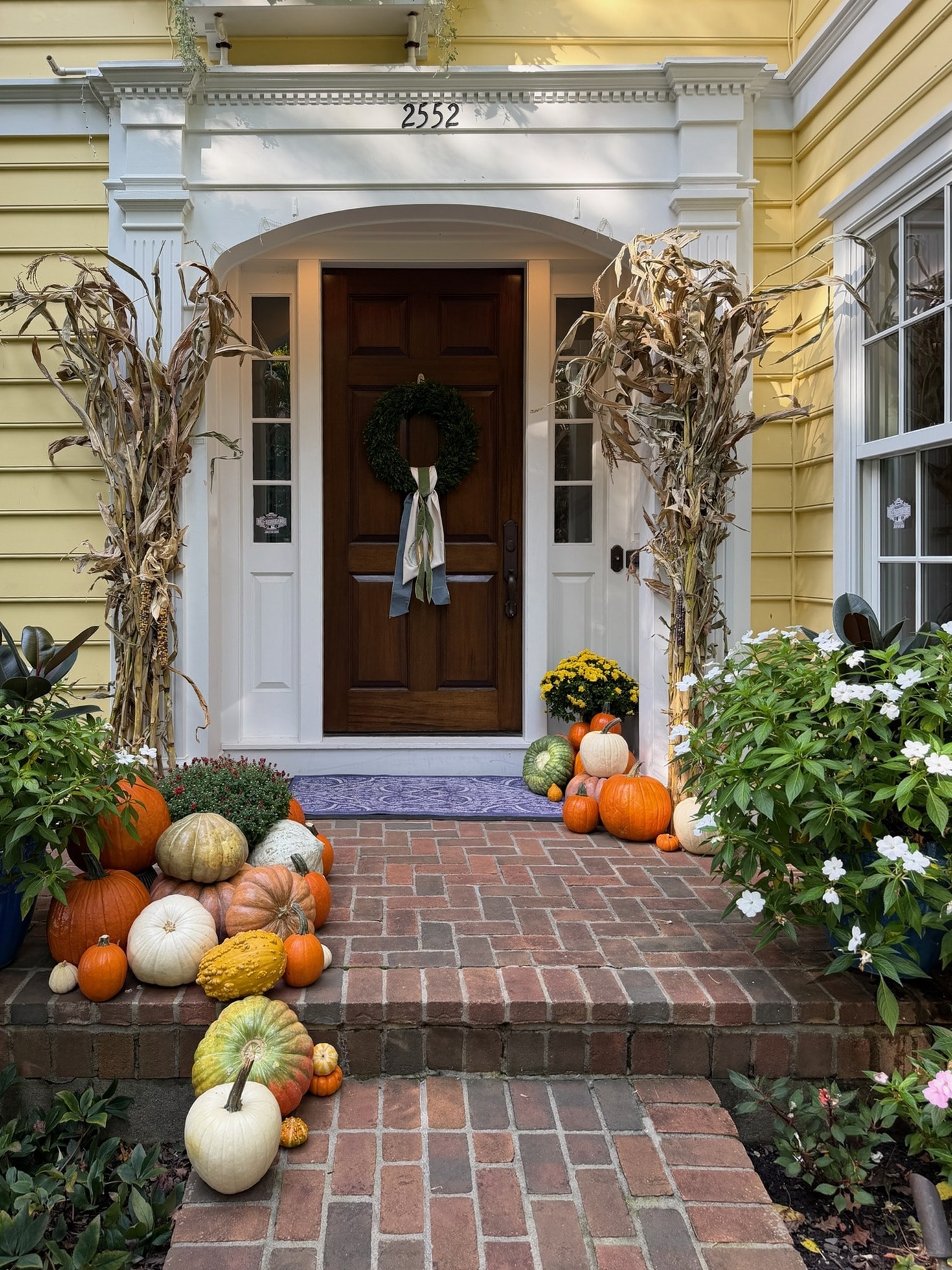 Yellow house with a wood front door framed by cornstalks, with pumpkins, gourds and mums arranged on the brick porch and steps