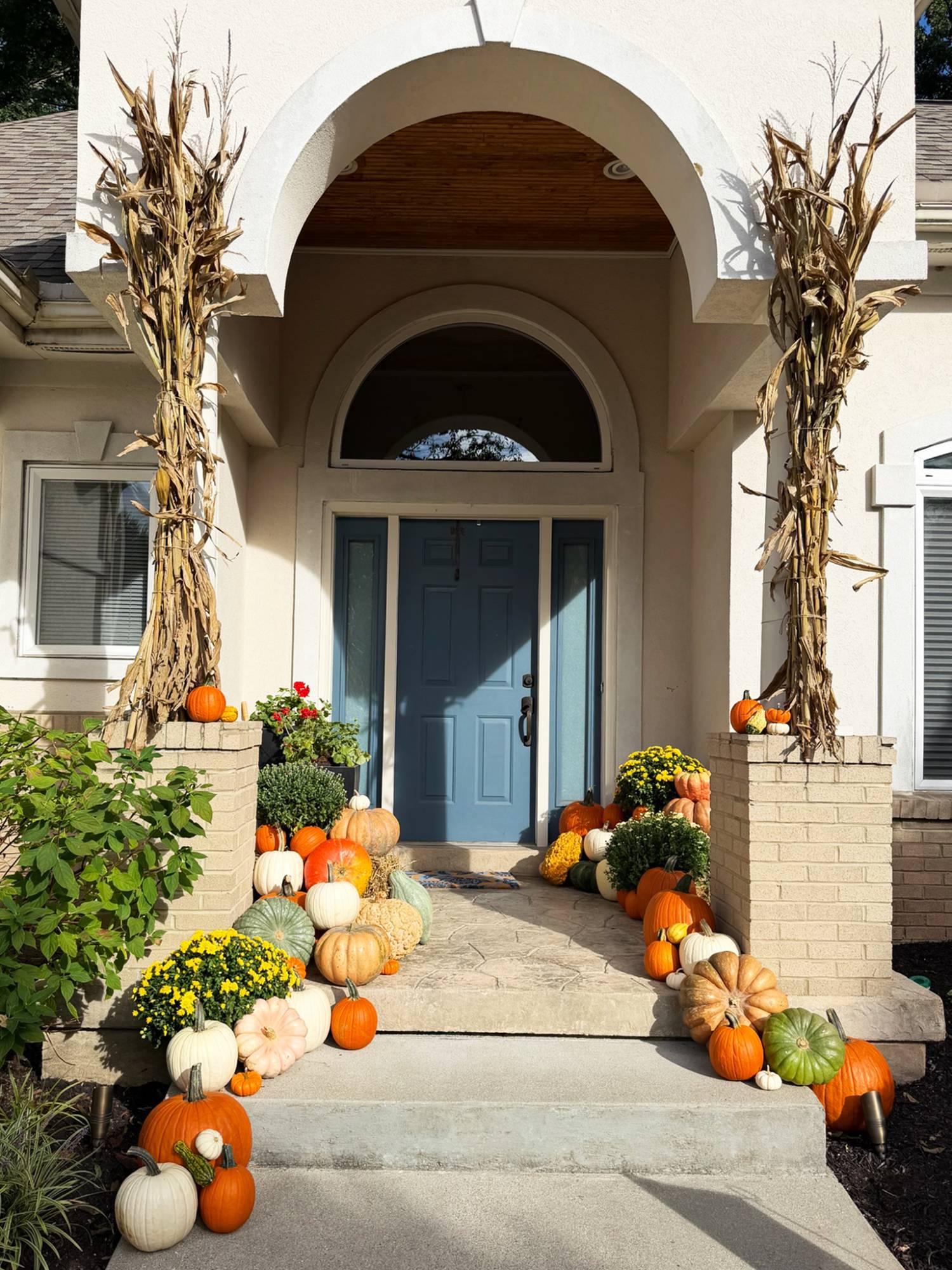 Stucco entrance with a blue front door, cornstalks tied to both brick pillars and pumpkins, gourds and mums grouped on the stoop and down the steps