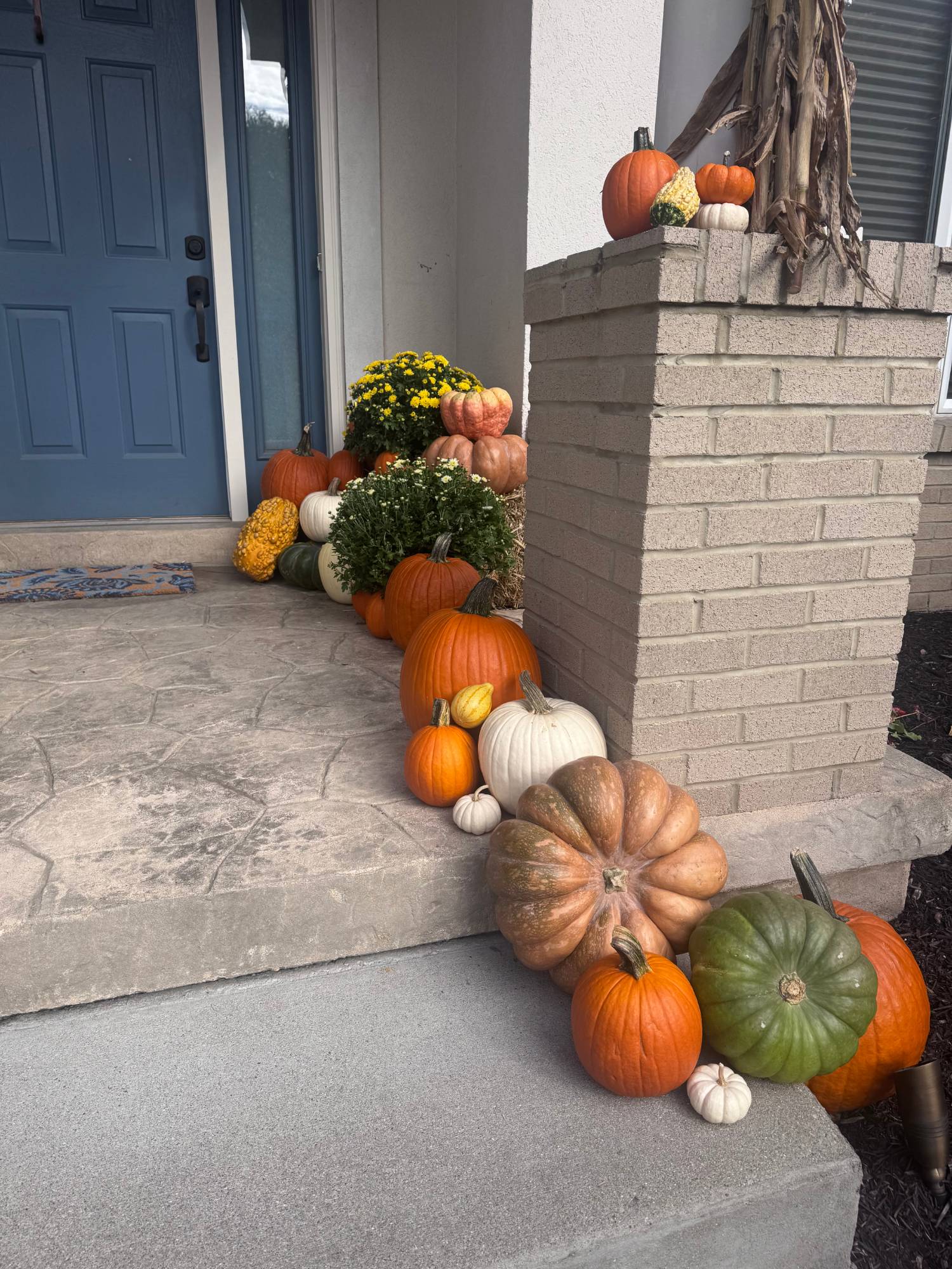 Heirloom, white and green pumpkins stepping down the stoop beside a brick pillar, with mums and a straw bale by the blue front door