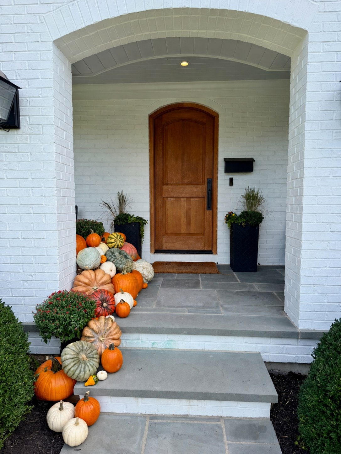 White brick entrance with an arched wood door and pumpkins and mums cascading down the left side of the stone steps