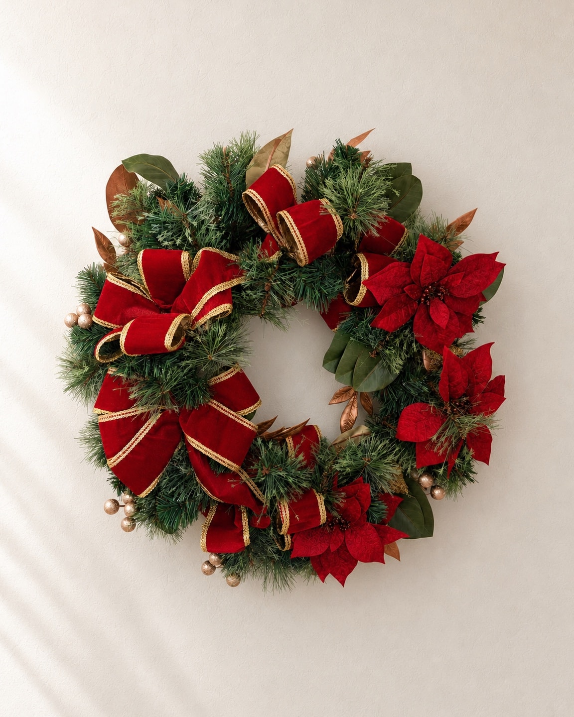 A wreath of fir and magnolia leaves with loops of red velvet ribbon, red poinsettia blooms and gold berries, on a pale plaster wall